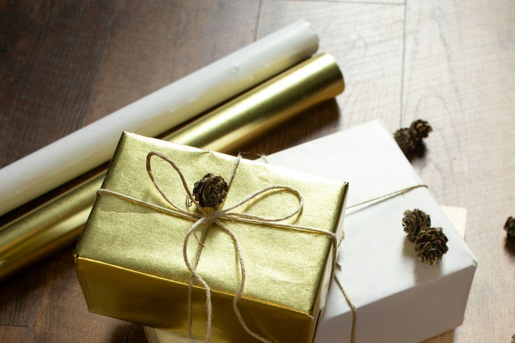 Two gifts wrapped in gold and white paper and tied with twine, with rolls of wrapping paper on a wooden floor
